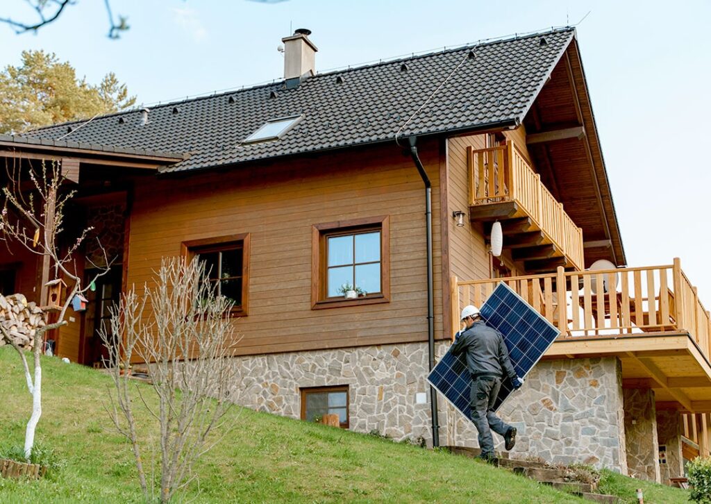 Ihre neue Solaranlage von Paul Energie - Ein Mann trägt ein Solarpanel vor einem Haus am grünen Hang
