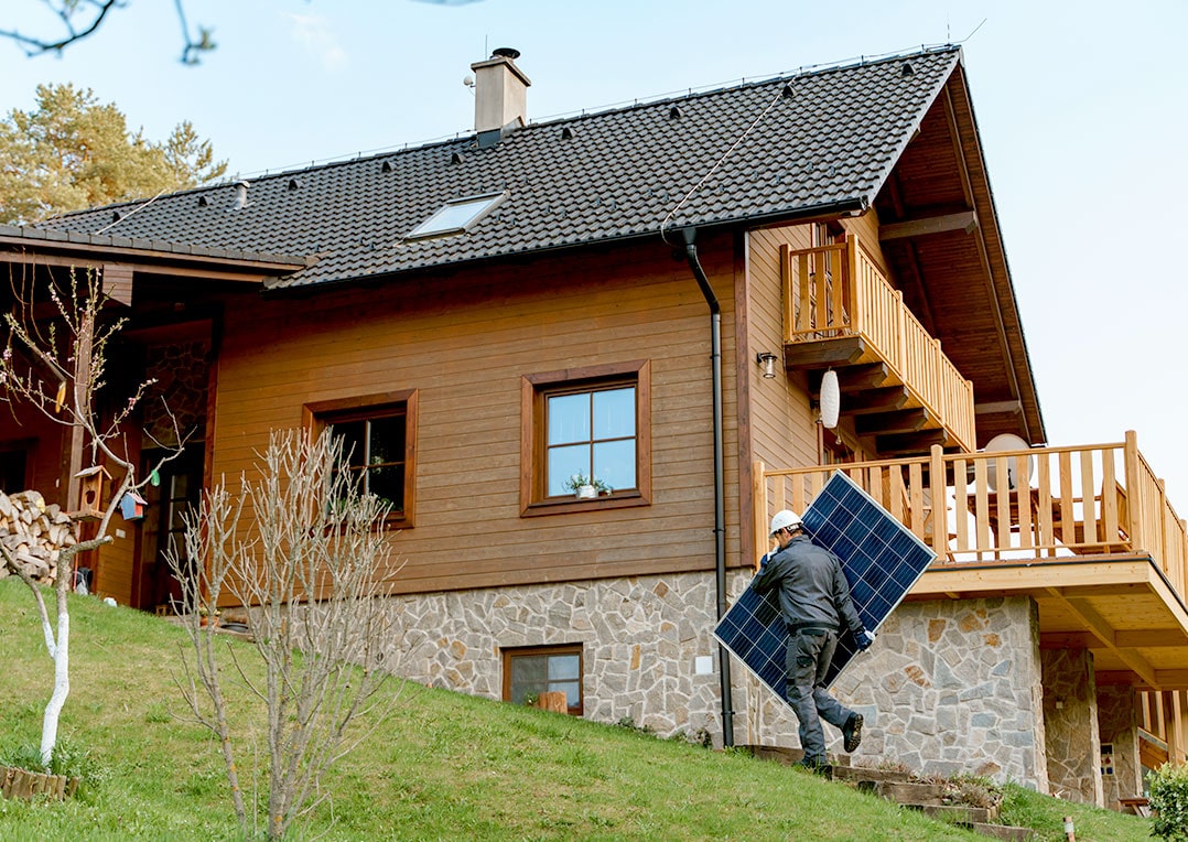 Ein Mann trägt ein Solarpanel vor einem Haus am grünen Hang – Ihre neue Solaranlage von Paul Energie